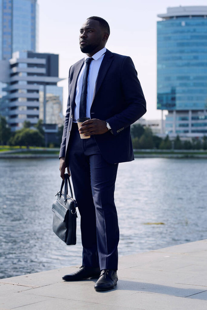 Junger erfolgreicher afrikanischer Geschäftsmann mit Handtasche bei einem Drink am Flussufer, während er an einem sonnigen Tag in der Stadt steht - Foto, Bild