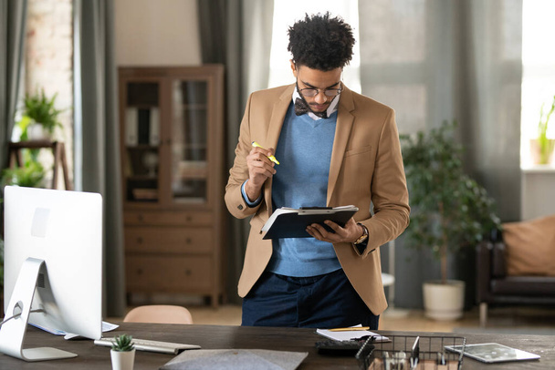 Gerente de marketing negro joven ocupado serio en chaqueta y suéter de pie en la mesa de madera y trabajando con papeles en la oficina en el hogar - Foto, Imagen