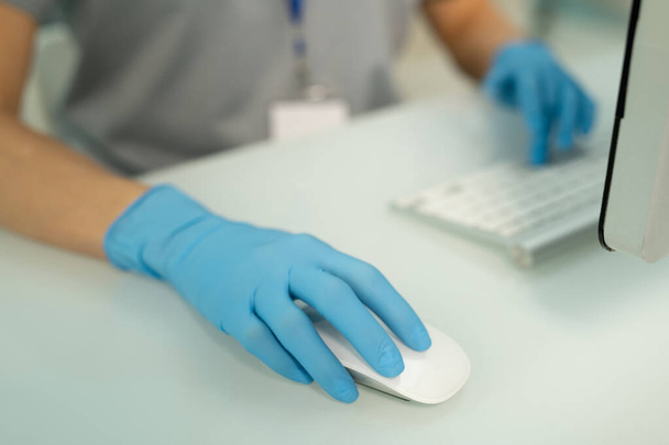 Крупним планом невизначена медсестра в синіх рукавичках сидить за столом і використовує комп'ютерну мишу при заповненні онлайн записів пацієнтів
 - Фото, зображення