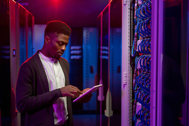 Серйозний молодий афроамериканський інженер даних стоїть у відкритому кабінеті сервера стійки і використовує планшет під час створення системи в центрі обробки даних. - Фото, зображення