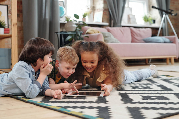 Grupo de niños felices tumbados en el suelo y jugando en tableta digital juntos en la habitación en casa - Foto, imagen