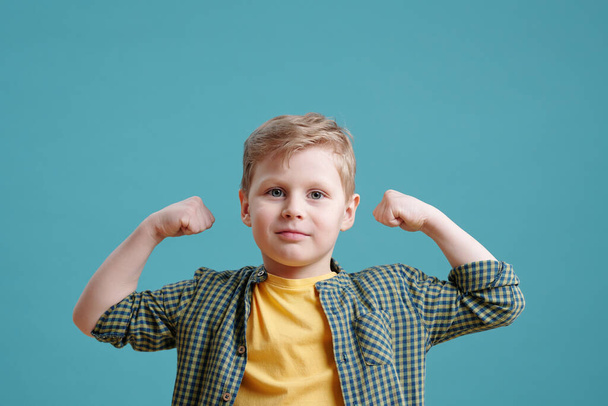 Portrait of cute little boy with blonde hair looking at camera and showing his power against the blue background - Foto, Imagen