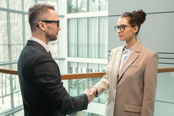 Handsome young businessman in suit standing in modern hall and shaking hand of beautiful lady while greeting her - 写真・画像