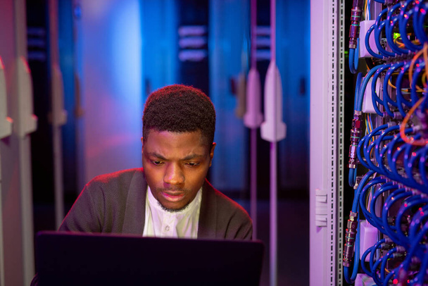 Απασχολημένος νεαρός μαύρος μηχανικός πληροφορικής που χρησιμοποιεί σύγχρονο φορητό υπολογιστή στο δωμάτιο διακομιστή ενώ διαχειρίζεται το δίκτυο υπολογιστών - Φωτογραφία, εικόνα