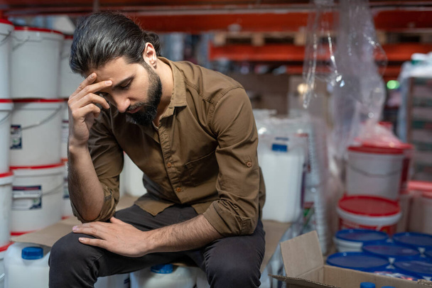 Puzzled young mixed race warehouse manager with beard sitting on canisters covered with cardboard and leaning head on hand - Photo, Image