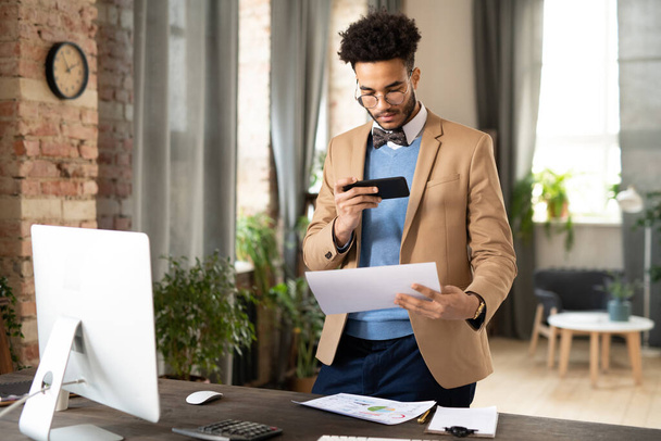 Drukke jonge gemengde ras werknemer in stijlvolle jas staan aan het bureau en het fotograferen van documenten op de smartphone in het kantoor - Foto, afbeelding