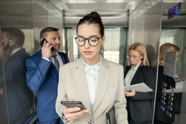 Asansörde çalışan ofis çalışanları: gözlüklü esmer kadın avukat telefonla mesaj atıyor. - Fotoğraf, Görsel
