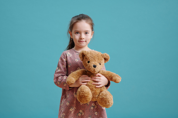 Porträt eines kleinen Mädchens mit Teddybär, das vor blauem Hintergrund in die Kamera blickt - Foto, Bild