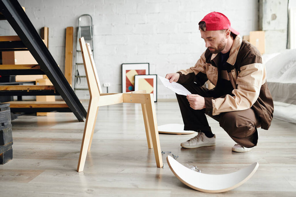 Junger Möbelmontagespezialist steht auf Kniebeugen und blättert bei der Montage eines neuen Holzstuhls in Anleitungen auf Papier - Foto, Bild