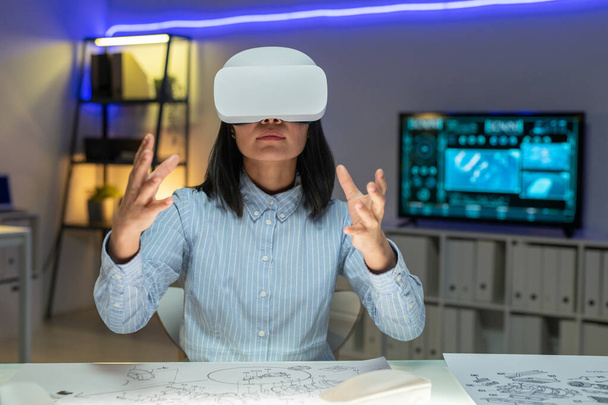 Mujer asiática joven concentrada en camisa formal sentada en el escritorio con bocetos y trabajando en el diseño 3D del coche en gafas VR - Foto, Imagen