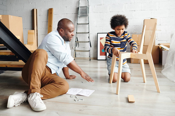 Genç Afrikalı bir adam, küçük şirin oğluna ahşap sandalyeyi nasıl monte edeceğini öğretirken ikisi de yeni dairelerinde ya da evlerinin zemininde oturuyorlar. - Fotoğraf, Görsel