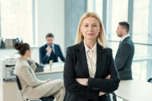 Retrato de contenido señora madura de pelo rubio en chaqueta negra de pie con los brazos cruzados en la oficina de negocios con colegas de fondo - Foto, Imagen