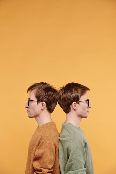Profile view of serious young redhead tweens in eyeglasses standing back to back against orange background - Photo, Image