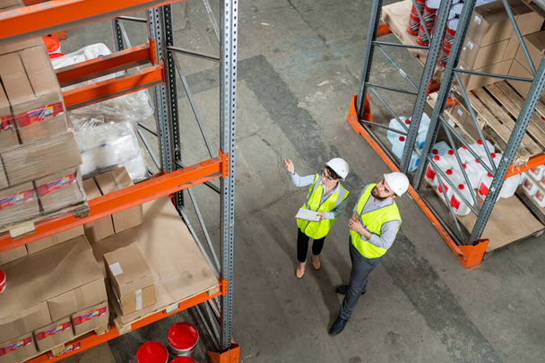 Directly above view of coworkers in green vests and hardhats discussing new supply at warehouse while looking at boxes on shelves - Photo, Image