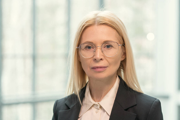 Portrait of serious beautiful mature executive in round glasses standing against window of office - Valokuva, kuva