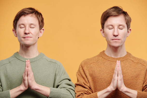 Ruhige, entspannte junge Tweens in Pullovern, die Hände in Namaste-Gesten halten und mit geschlossenen Augen vor orangefarbenem Hintergrund meditieren - Foto, Bild