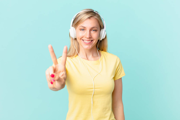 young blonde woman smiling and looking happy, gesturing victory or peace and listening music. - Фото, изображение