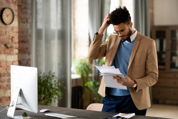 Confused young mixed race man in glasses and jacket standing at desk with computer and scratching head while reading papers - Photo, Image