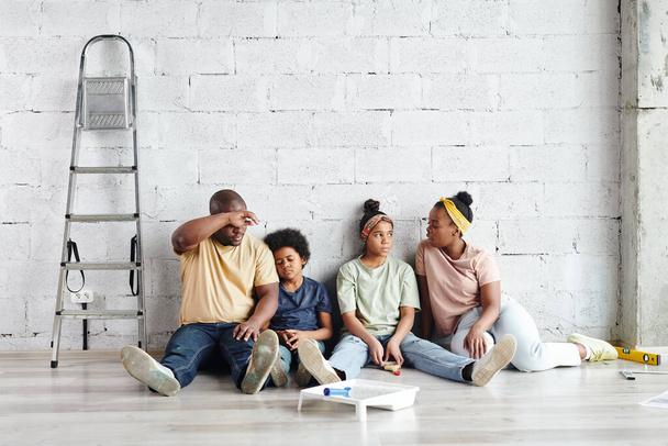 Κουρασμένη οικογένεια τεσσάρων Αφρικανών που κάθεται απέναντι σε λευκό βαμμένο τοίχο στο πάτωμα του σαλονιού μετά τις εργασίες ανακαίνισης του σπιτιού και έχουν ξεκουραστεί μαζί - Φωτογραφία, εικόνα