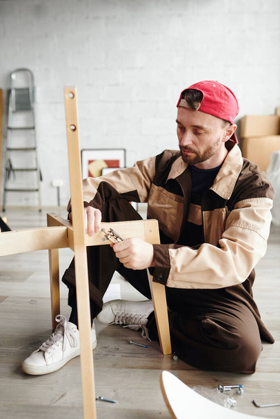 Serious young furniture assemblage specialist in workwear sitting on the floor and fixing parts of chair while assembling it in new flat or studio - Foto, Imagen