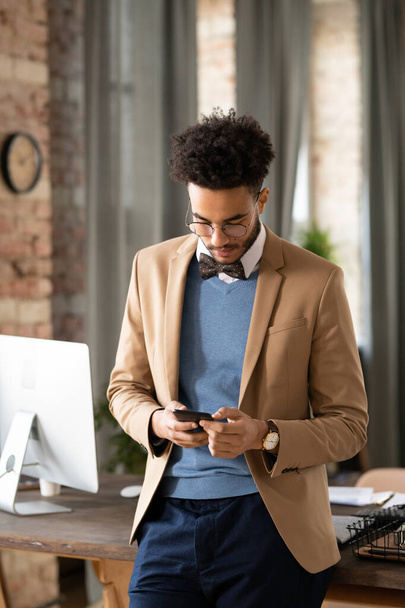 Concentrado jovem homem negro em gravata borboleta de pé na mesa do escritório e verificando mensagens de telefone - Foto, Imagem