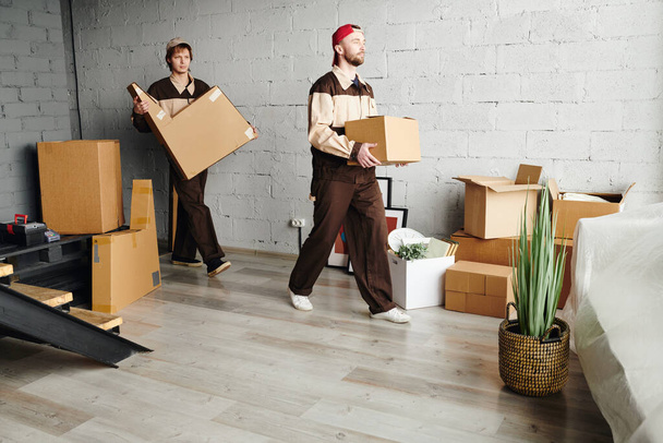 Twee jonge laders in werkkleding die verpakte kartonnen dozen dragen terwijl ze helpen om pakketten te leveren aan nieuwe flat, huis of studio - Foto, afbeelding