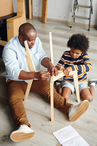 Joven hombre de etnia africana enseñando a su pequeño y lindo hijo cómo montar muebles nuevos mientras está sentado en el piso de un nuevo piso o casa - Foto, imagen