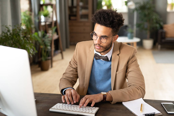 Vážný mladý afro americký podnikatel v brýlích a náramkových hodinkách sedí u stolu a psaní na počítači v útulné kanceláři - Fotografie, Obrázek