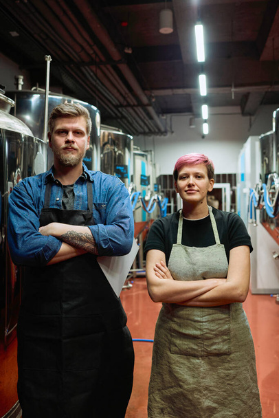 Jóvenes trabajadores serios de la fábrica de cerveza contemporánea cruzando los brazos por el pecho mientras están de pie uno al lado del otro delante de la cámara - Foto, Imagen