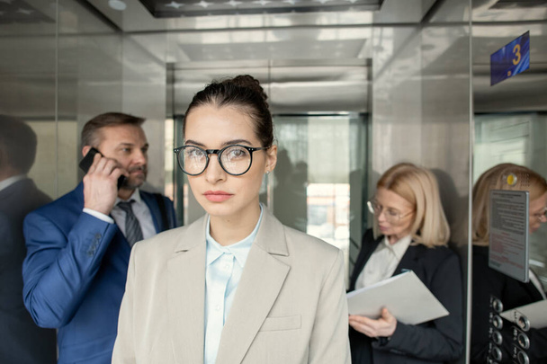 Портрет серйозної привабливої бізнес-леді, що стоїть в ліфті з зайнятими людьми на задньому плані
 - Фото, зображення