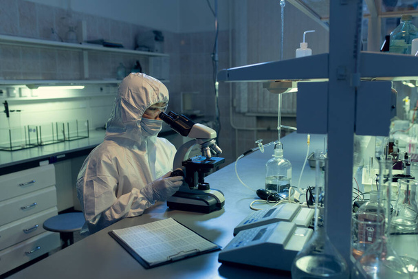 Científico en ropa de trabajo protectora que examina la bacteria a través del microscopio en la mesa del laboratorio - Foto, Imagen
