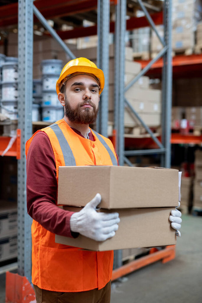 Portrait of content confident young depository mover in hardhat and reflective vest standing with boxes against warehouse frame - Fotó, kép