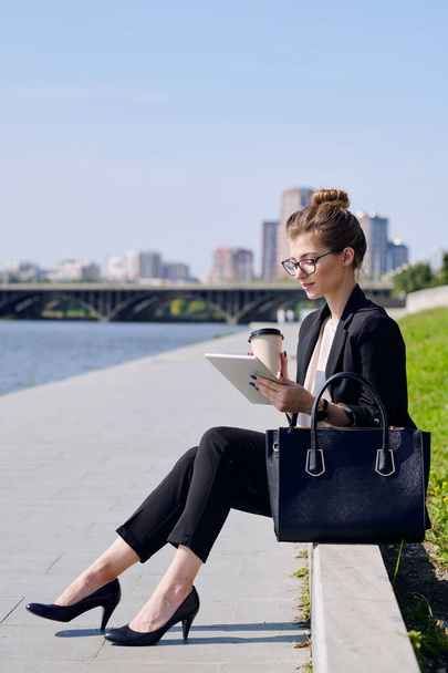 Giovane donna bionda impegnata in abbigliamento formale che beve e usa tablet mentre si siede di fronte al fiume in ambiente urbano nella giornata di sole - Foto, immagini