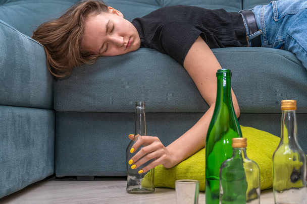 Κουρασμένη νεαρή μελαχρινή γυναίκα ξαπλωμένη στον καναπέ μετά από ένα πάρτι στο σπίτι, δίπλα σε άδεια μπουκάλια στο πάτωμα. - Φωτογραφία, εικόνα