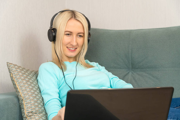 Close up smiling young blonde woman in bright clothes is lying on the sofa with a laptop in large headphones - Foto, imagen