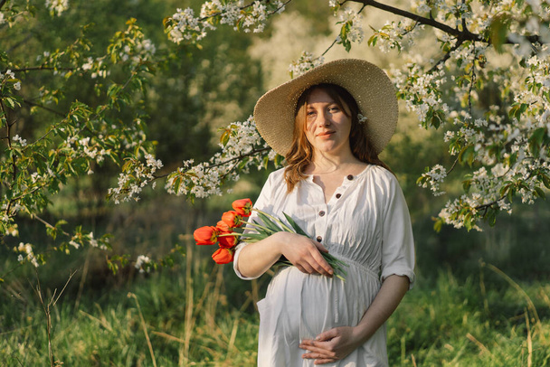 Retrato de primavera de uma mulher grávida. Uma bela jovem grávida em um vestido branco caminha no jardim da primavera. Feliz gravidez. Primavera - Foto, Imagem