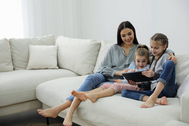 Mother and daughters with tablet sitting on comfortable sofa in living room - Zdjęcie, obraz