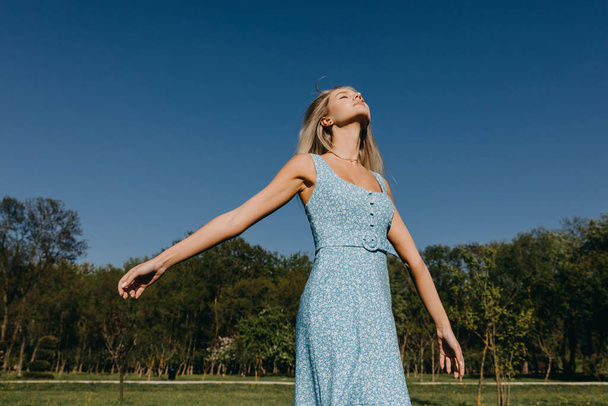 Молодая женщина с длинными светлыми волосами, в синем платье, наслаждается хорошей погодой, на открытом воздухе. - Фото, изображение