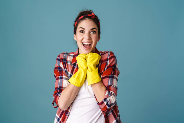Smiling excited attractive young woman in apron and rubber gloves standing isolated over blue background - Zdjęcie, obraz