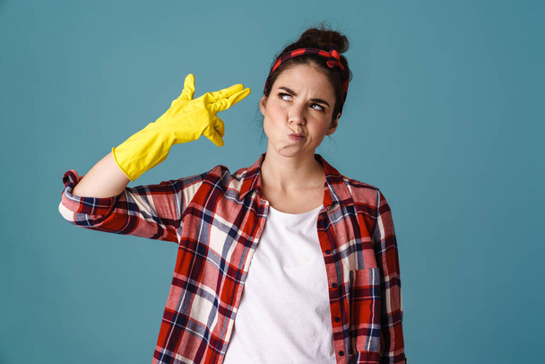 Tired beautiful girl in gloves grimacing and making gun gesture isolated over blue background - Фото, изображение