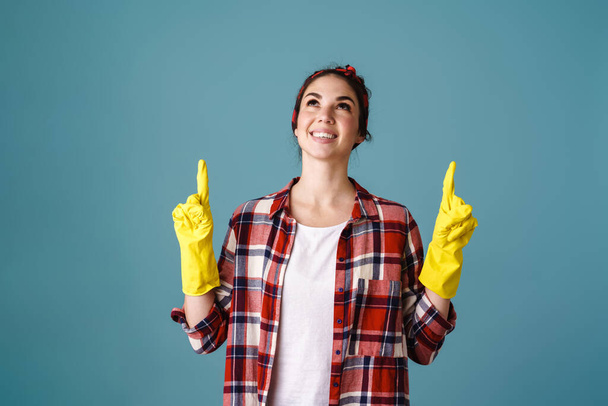 Happy beautiful girl in gloves smiling and pointing fingers upward isolated over blue background - 写真・画像