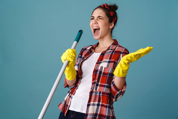 Happy cheerful young woman in apron and rubber gloves holding mop isolated over blue background - Фото, изображение