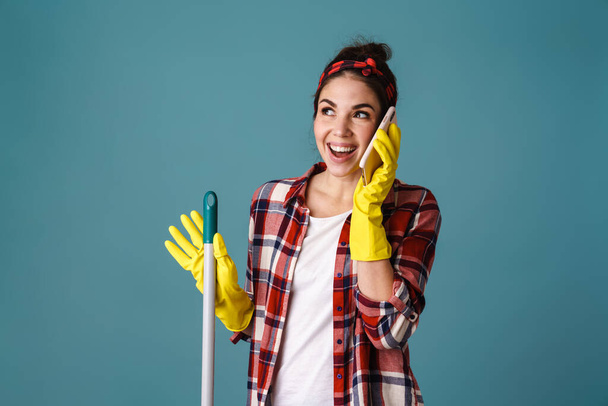 Excited attractive woman in rubber gloves and apron using mobile phone isolated over blue background - Φωτογραφία, εικόνα