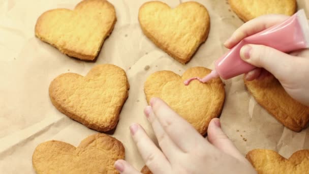 nainen koristaa sydämen muotoisia evästeitä vaaleanpunaisella kuorrutuksella, lähikuva, leivinprosessi, romanttinen käsite - Materiaali, video
