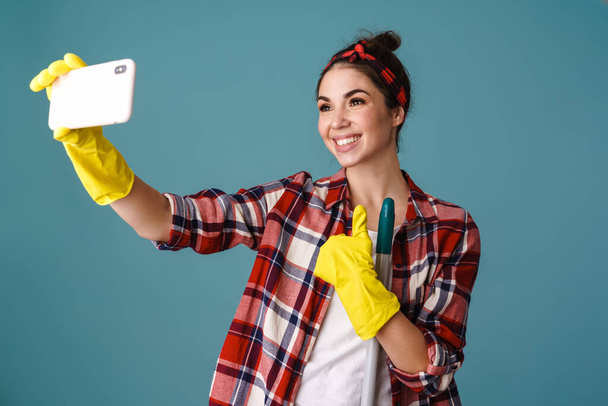 Smiling beautiful girl in gloves with mop taking selfie on cellphone isolated over blue background - Фото, зображення