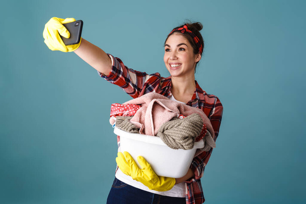 Happy girl in gloves with dirty clothes taking selfie on cellphone isolated over blue background - Foto, Bild