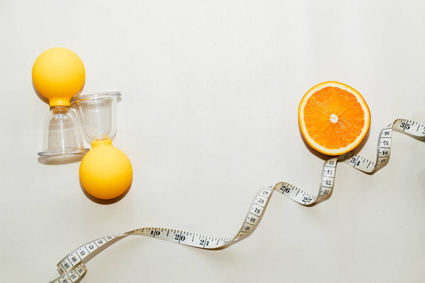 Вакуумный массаж антицеллюлитных чашек на сером фоне с апельсином и сантиметром. Самомассаж для здоровой кожи. - Фото, изображение