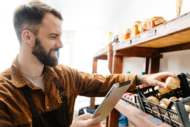 Smiling bearded seller man using tablet computer while working in local eco shop - Fotó, kép