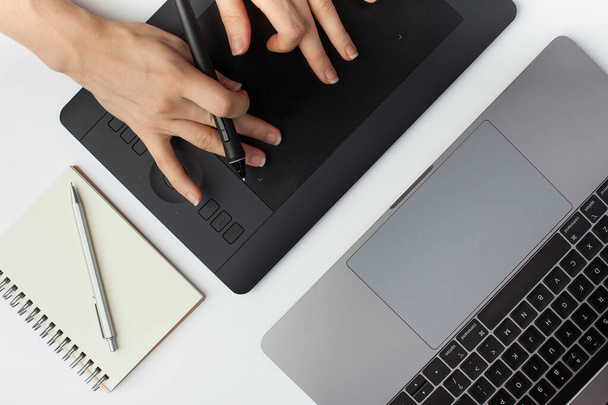 Foto de vista superior de uma mão feminina ampliando em um tablet de design gráfico preto, caneta, bloco de notas e um laptop em um fundo branco. - Foto, Imagem