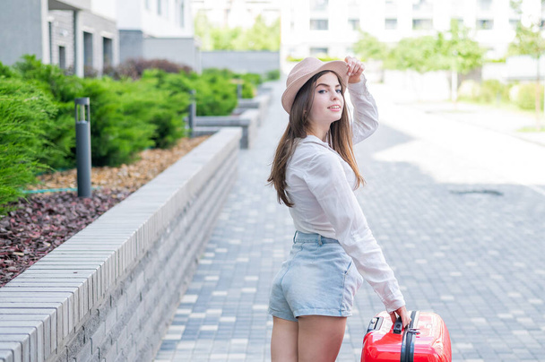 Young caucasian woman in a hat and shorts stands on the street with a red suitcase. - Photo, image